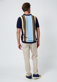 Man staat met zijn rug naar de camera, gekleed in beige broek, een marineblauw en lichtblauw gestreept T-shirt met korte mouwen, en navy sneakers met mosterdkleurige accenten.