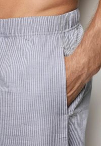 La mano de un hombre descansando en el bolsillo de unos pantalones de cintura elástica de rayas verticales en gris claro y blanco.