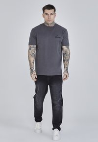 Grijze katoenen T-shirt met ronde hals en korte mouwen, voorzien van een zwart geborduurd logo. Draagt met donkere jeans en witte sneakers.