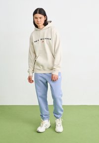 Beige Kapuzenpullover mit "NEW BALANCE"-Logo, kombiniert mit hellblauen Sweatpants. Modell trägt weiße Sneaker mit reflektierenden Akzenten. Schlichter, lässiger Look.