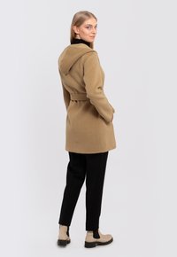 Knee-length coat in light brown wool, featuring a hood, belt at the waist, and side pockets, paired with black trousers and beige ankle boots.