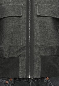 Cardigan gris avec fermeture éclair à l'avant, deux poches plaquées, ourlet et poignets côtelés. Fabriqué en tissu doux avec une finition texturée.