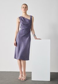 Lila linneklänning med asymmetrisk halsringning, draperad detaljering och knälång fåll. Matchad med vita sandaler med remmar och silverarmband.