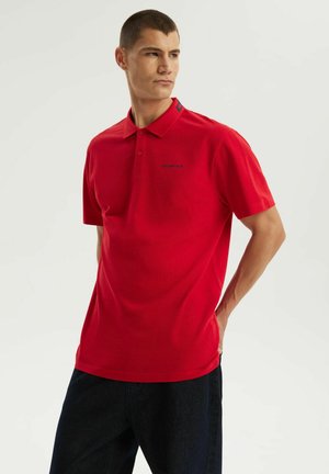 Młody mężczyzna w czerwonej koszulce polo z krótkim rękawem i ciemnych spodniach stoi z rękami za plecami, patrząc na bok.