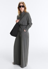 Femme portant un costume gris à rayures fines et pantalon large, lunettes de soleil noires, tenant un sac noir sur l'épaule, debout devant un fond blanc.