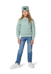 Light green knitted sweater with a textured logo, paired with blue denim jeans and white sneakers. The model also wears a matching beanie.