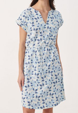 Femme portant une robe blanche longueur genou avec des motifs floraux bleus, manches courtes, col en V et taille à cordon.