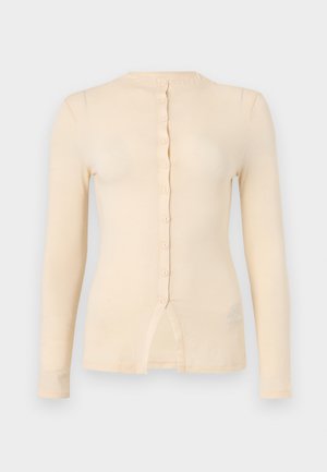 Μπεζ εφαρμοστό μακρυμάνικο τοπ με στρογγυλή λαιμόκοψη, κούμπωμα μπροστά και μικρή σχισμή στο κέντρο του κάτω μέρους.