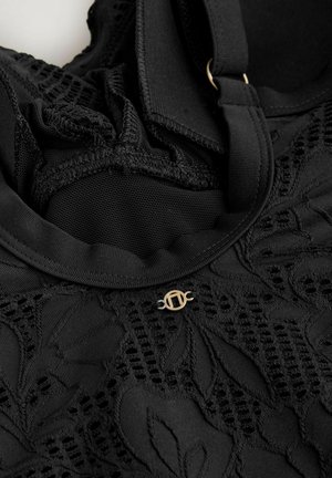 Svart textil med intrikata lövmönster och detaljer i mesh, med en guldfärgad logotyp som är fäst vid remmen. Mjukt, texturerat tyg.