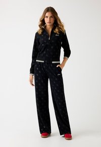 Zwarte velours trainingsoutfit met een ritsjack en losse broek, voorzien van een subtiel patroon en witte gestreepte accenten. Rode instap schoenen maken de look compleet.