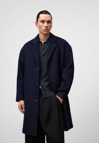 Marineblauer Wollmischungs-Mantel mit großen Revers, einreihigem Design und drei Knöpfen, kombiniert mit einem dunklen Poloshirt und maßgeschneiderten Hosen.