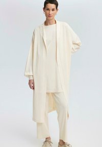 Completo tre pezzi color beige chiaro composto da un cardigan lungo e aperto, una semplice maglietta e pantaloni a sigaretta, tutti realizzati in un tessuto morbido e liscio.