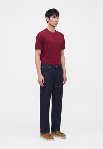 Hombre de pie con camiseta roja de manga corta, vaqueros azul oscuro y zapatillas marrones sobre un fondo claro liso.