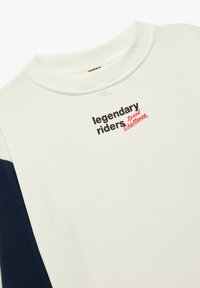 Biała bluza z granatowymi rękawami, z czarnym i czerwonym napisem: "legendarny jeźdźcy" oraz "Wyzwanie Prędkości". Gładka faktura, fason z okrągłym dekoltem.