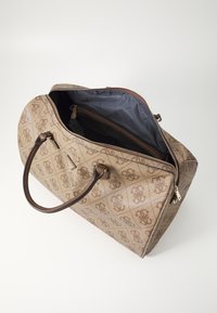 Sac de sport beige avec un extérieur à motifs, des accents en cuir marron, deux poignées et une ouverture zippée qui révèle un intérieur sombre.