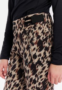 Leopardprint skibroek in beige, bruin en zwart; heeft een platte tailleband, voorzakken en verstelbare broekspijpen met koordjes.