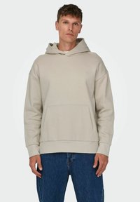 Hoodie beige chiaro realizzato in tessuto morbido, presenta una tasca a canguro, maniche larghe e un cappuccio con cordino. Abbinato a jeans di denim scuro.