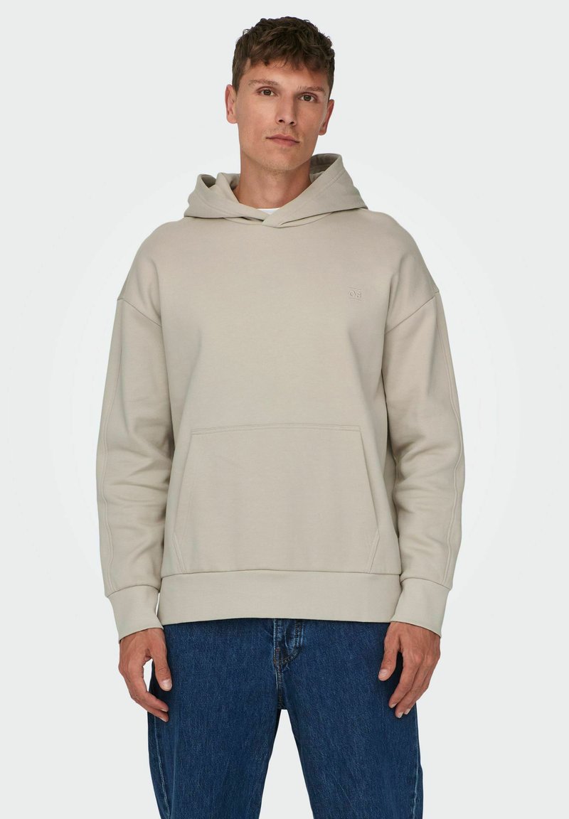 Hoodie beige chiaro realizzato in tessuto morbido, presenta una tasca a canguro, maniche larghe e un cappuccio con cordino. Abbinato a jeans di denim scuro.