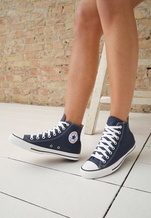 Nogi noszące granatowe wysokie trampki Converse z białymi sznurówkami, stojące na białej drewnianej podłodze na tle ceglastej ściany.