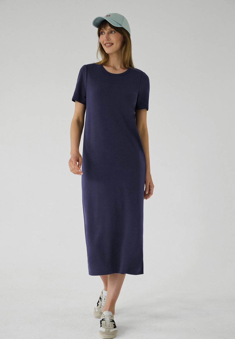 Femme portant une robe midi bleu marine, une casquette de baseball bleu clair et des baskets, debout devant un fond uni gris clair.