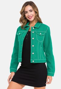 Grüne Jeansjacke mit weißen Nähten, zwei Brusttaschen und silbernen Knöpfen, kombiniert mit einem schwarzen figurbetonten Kleid.