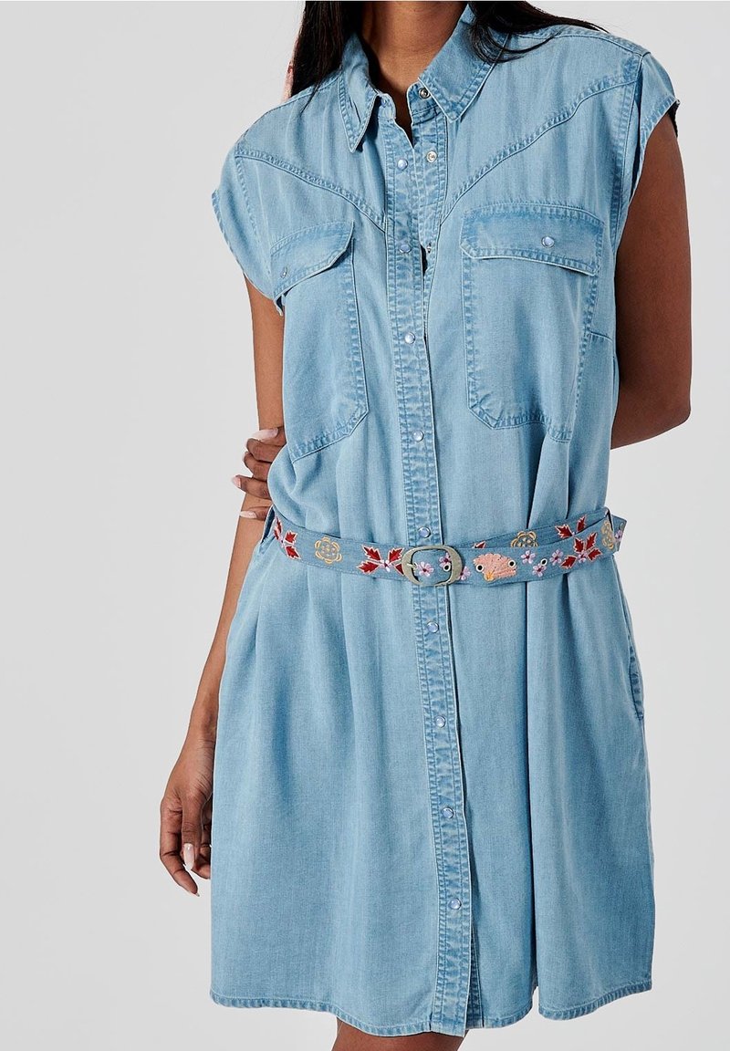 Robe chemise en denim bleu clair avec des manches courtes, deux poches poitrine, une fermeture à boutons sur le devant, et une ceinture brodée avec des accents floraux colorés.