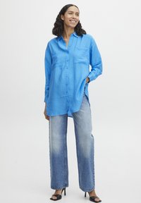 Camicia blu con bottoni, finitura testurizzata, due tasche e maniche lunghe, abbinata a jeans in denim sbiadito a gamba larga e tacchi neri.