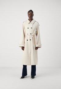 Kremfarget trenchcoat med doble svarte knapper, vide ermer, belte og skulderepauleter, kombinert med mørke vide jeans og svarte hæler.