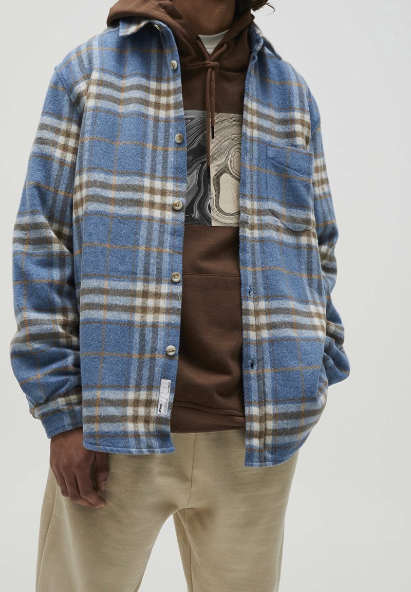 Blue flannel shirt with a plaid pattern, featuring beige and gray lines. Button closure, patch pocket, layered over a brown hoodie.