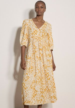 Kobieta w żółto-białej maxi sukience w wzory z rękawami o długości do łokcia z bufkami i dekoltem w serek, stojąca na jednolitym tle.