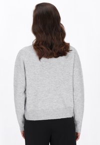 Vrouw met golvend bruin haar, gekleed in een lichtgrijze trui en zwarte broek, staand met haar rug naar de camera tegen een witte achtergrond.