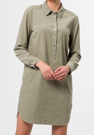 Langärmliges olivgrünes Cordhemdkleid mit silbernen Knöpfen und Kragen, getragen von einer Person mit verschränkten Händen auf der Taille.