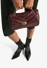 Burgundy läderhandväska med chevrondesign, guldhårdvara och ett lås. Bärs med svarta byxor och mönstrade strumpor, med spetsiga klackar.