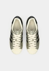 Pár olivovo zelených a čiernych tenisiek Adidas s bielymi špičkami a krémovými šnúrkami, zobrazené zhora na bielom pozadí.