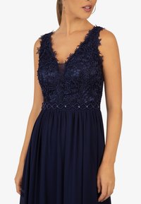Abito da sera blu navy con scollo a V profondo, corpino in pizzo floreale, dettagli in perline e gonna fluida realizzata in tessuto leggero.