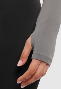 Graues Langarmshirt aus Stoff mit Daumenlochdetail, kombiniert mit schwarzem Material. Glatte Textur, taillierte Passform, die das Handgelenk betont.