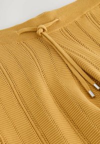 Tissu tricoté jaune avec des rayures verticales ajourées et un cordon de serrage noué avec des embouts en métal sur une ceinture côtelée.