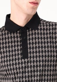 Mustra musta ja hall polo särk, millel on korduv muster tumedate aktsentidega, kolme nupuga takjaga ja must krae.