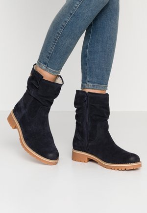 Granatowe zamszowe buty za kostkę z beżowymi gumowymi podeszwami, bocznymi zamkami błyskawicznymi i ozdobnymi paskami, noszone z niebieskimi obcisłymi jeansami.