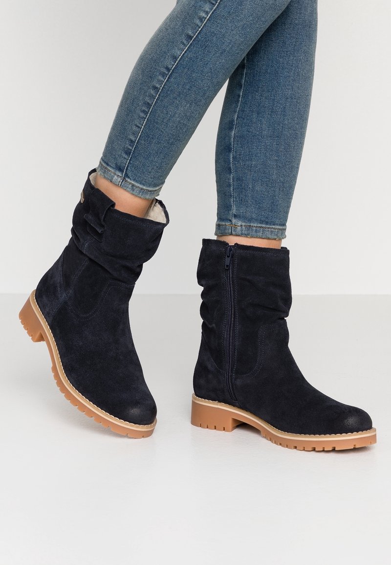 Bottes mi-mollet en daim bleu foncé avec semelles en caoutchouc couleur fauve, fermetures éclair latérales et brides décoratives, portées avec un jean skinny bleu.