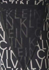 Czarna tkanina z wyraźnym, powtarzającym się białym tekstem w nowoczesnej czcionce. Projekt zawiera dekolt w kształcie litery V oraz teksturowane akcenty.