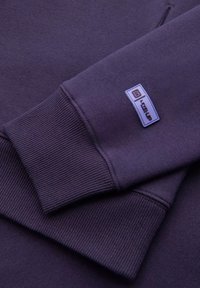 Sweatshirt violet avec des poignets côtelés et un écusson logo en forme de carré. Il présente une texture douce et une couleur uniforme, sans motifs.
