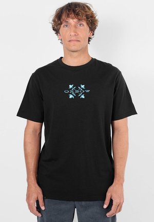 Homme aux cheveux bouclés portant un T-shirt noir avec un motif floral bleu et le texte "Oxbow" sur la poitrine, debout devant un fond uni.