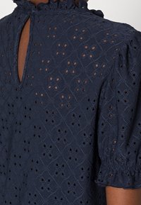 Mörkblå blus med en strukturerad tyg, broderade diamantmönster och utskärningar, korta puffärmar och en volangkrage.