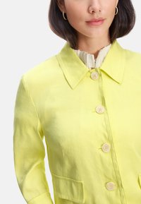 Yellow button-up jacket made of lightweight fabric, featuring a spread collar, front pockets, and natural finish with textured stitching.