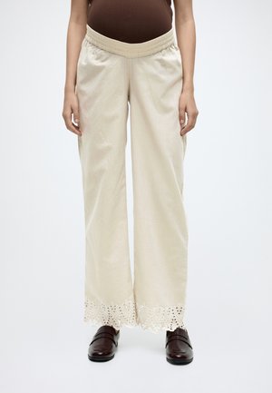 Beige wijde broek met elastische tailleband en kanten rand met oogjes, gedragen met donkerbruine loafers en een bruin shirt.