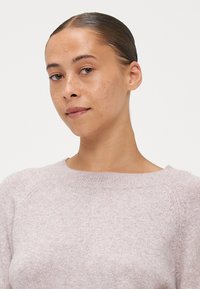 Sweater rosa claro com decote redondo, mangas raglan e tecido suave e texturizado. Close-up mostrando detalhes suaves e variações subtis.