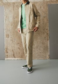Beige canvas jacket with black collar and front pockets, paired with beige pants and black sneakers featuring a green pattern.