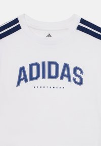 Biała bawełniana koszulka z granatowymi paskami na ramionach, z napisem "ADIDAS" dużymi niebieskimi literami oraz "SPORTSWEAR" mniejszym tekstem poniżej.