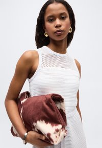 Γυναίκα που φοράει λευκό πλεκτό φόρεμα χωρίς μανίκια, κρατώντας μια διπλωμένη καφέ και λευκή κουβέρτα με σχέδιο σε απλό φόντο.